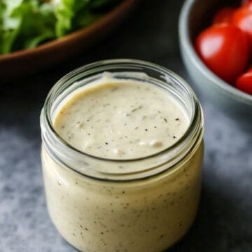 A jar of caesar dressing with a bowl of tomatoes and a bowl of salad in the background.