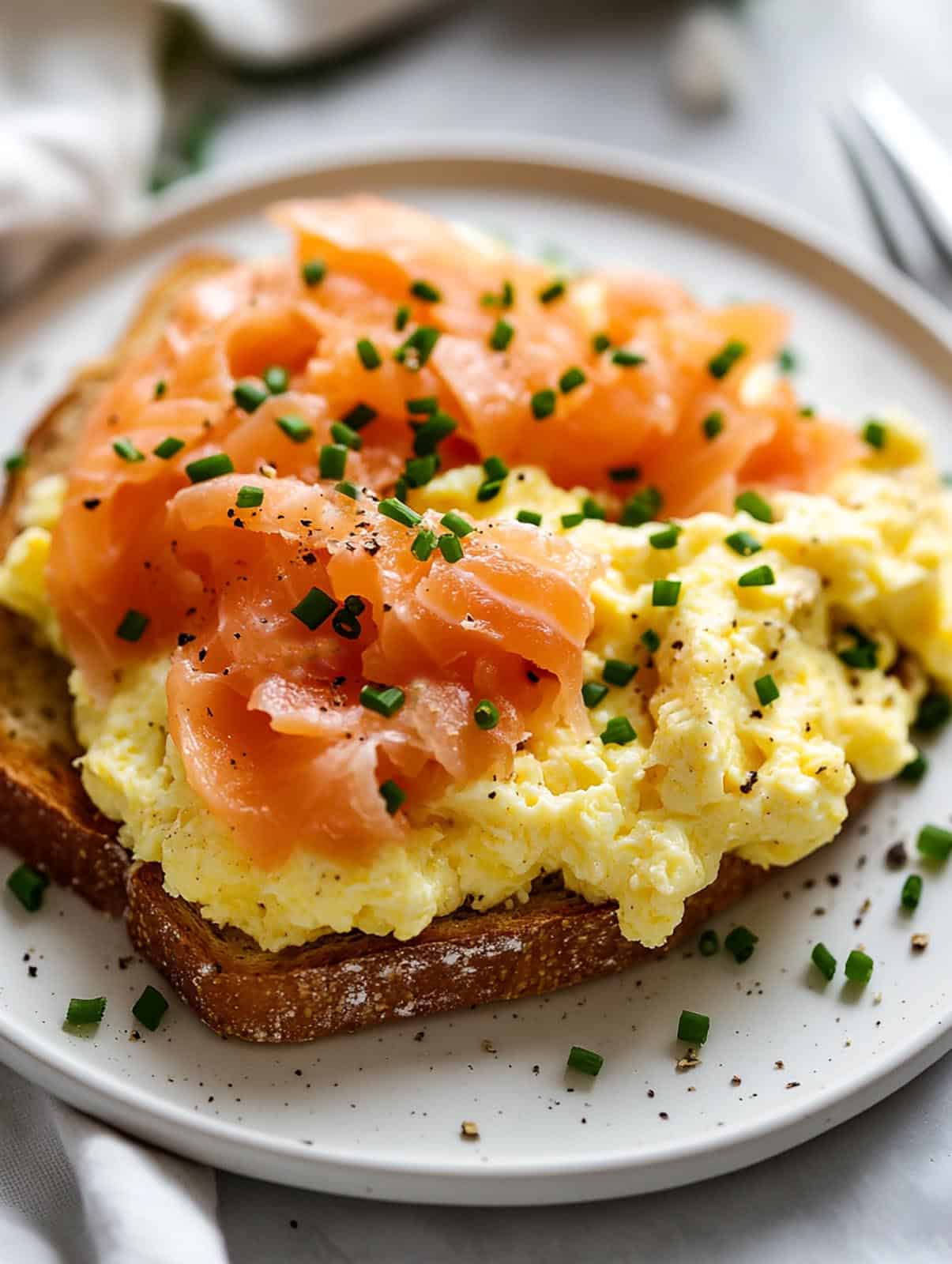 A white plate of scrambled eggs on toast with smoked salmon and chopped chives.