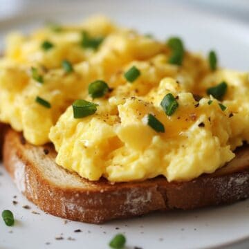 A close up image of scrambled eggs on toast topped with chives and on a white plate.