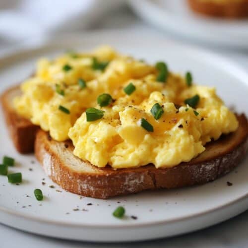 A close up image of a white plate with scrambled eggs on toast topped with sliced chives.