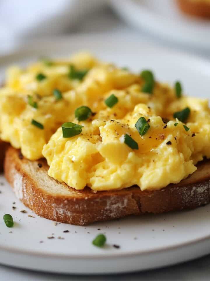 A close up image of a white plate with scrambled eggs on toast topped with sliced chives.