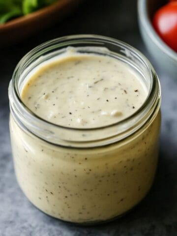 A jar of creamy caesar dressing on a grey table with a bowl of tomatoes in the background.