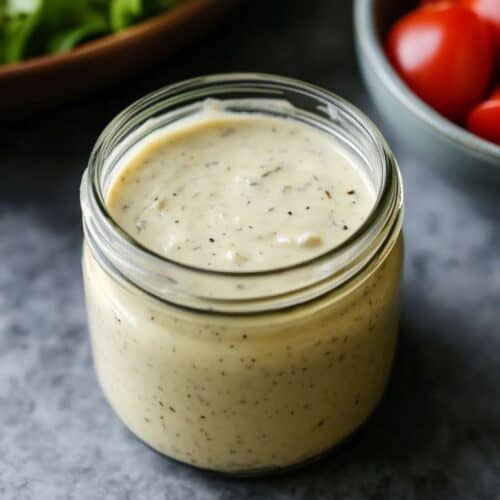 A jar of creamy caesar dressing on a grey table with a bowl of tomatoes in the background.