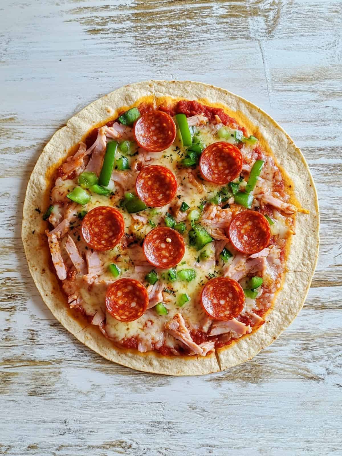 An image of a tortilla wrap pizza topped with pepperoni, melted cheese, chopped green veggies and turkey on a white table.