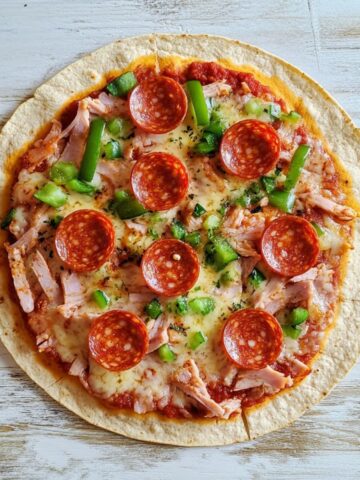 An image of a tortilla wrap pizza topped with pepperoni, melted cheese, chopped green veggies and turkey on a white table.