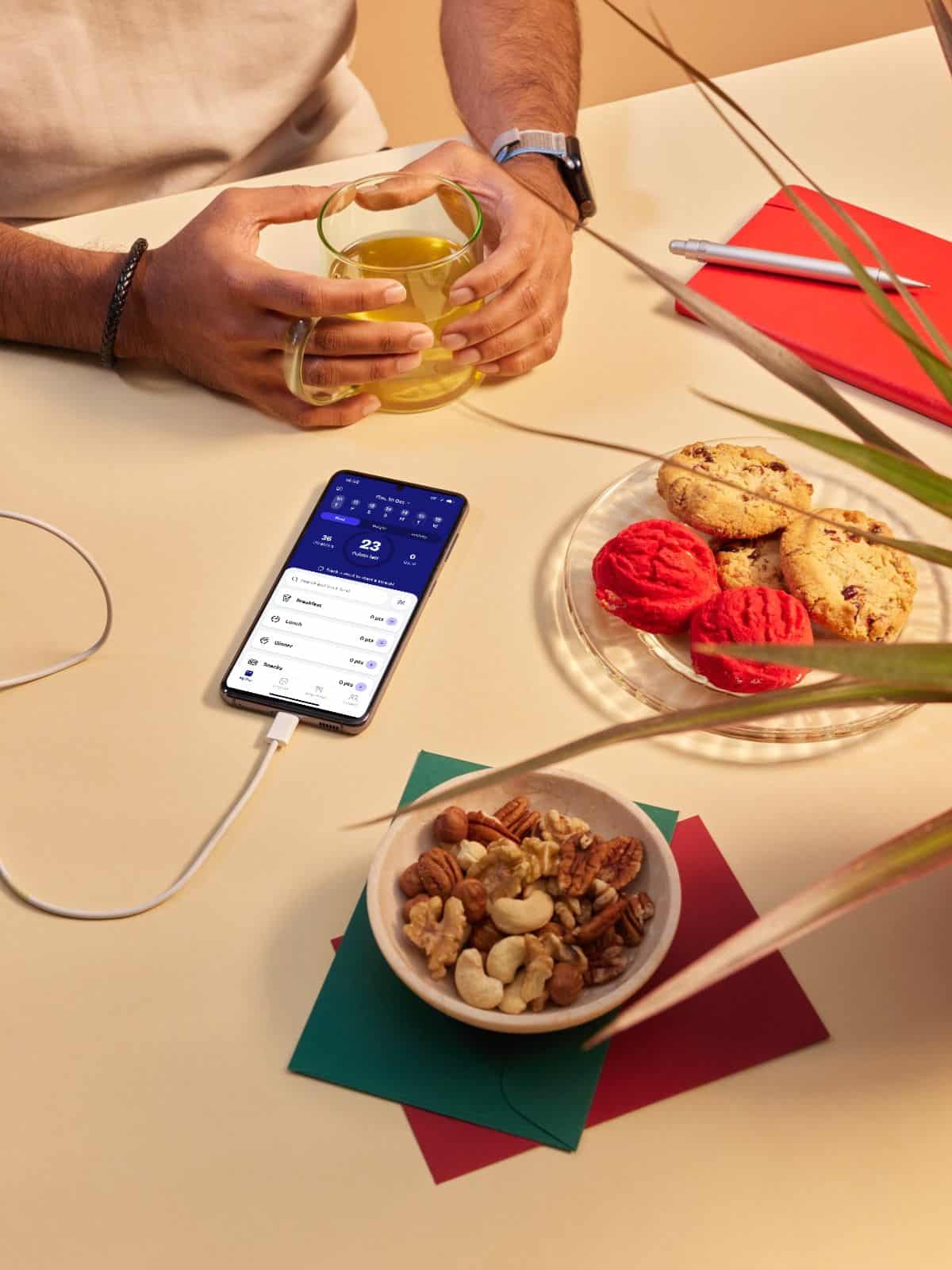 An image of a person sitting at a table with a bowl of nuts and a plate of cookies. Beside them is a phone charging and showing the WW App.