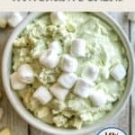 An image of watergate salad in a white bowl on a painted table. On the table are marshmallows and pistachio nibs. There is text overlay stating Weight Watchers Watergate Salad, Low in Points.