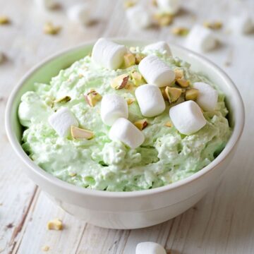 An image of watergate salad in a white bowl on a white painted table. On the table are marshmallows and pistachio nibs.