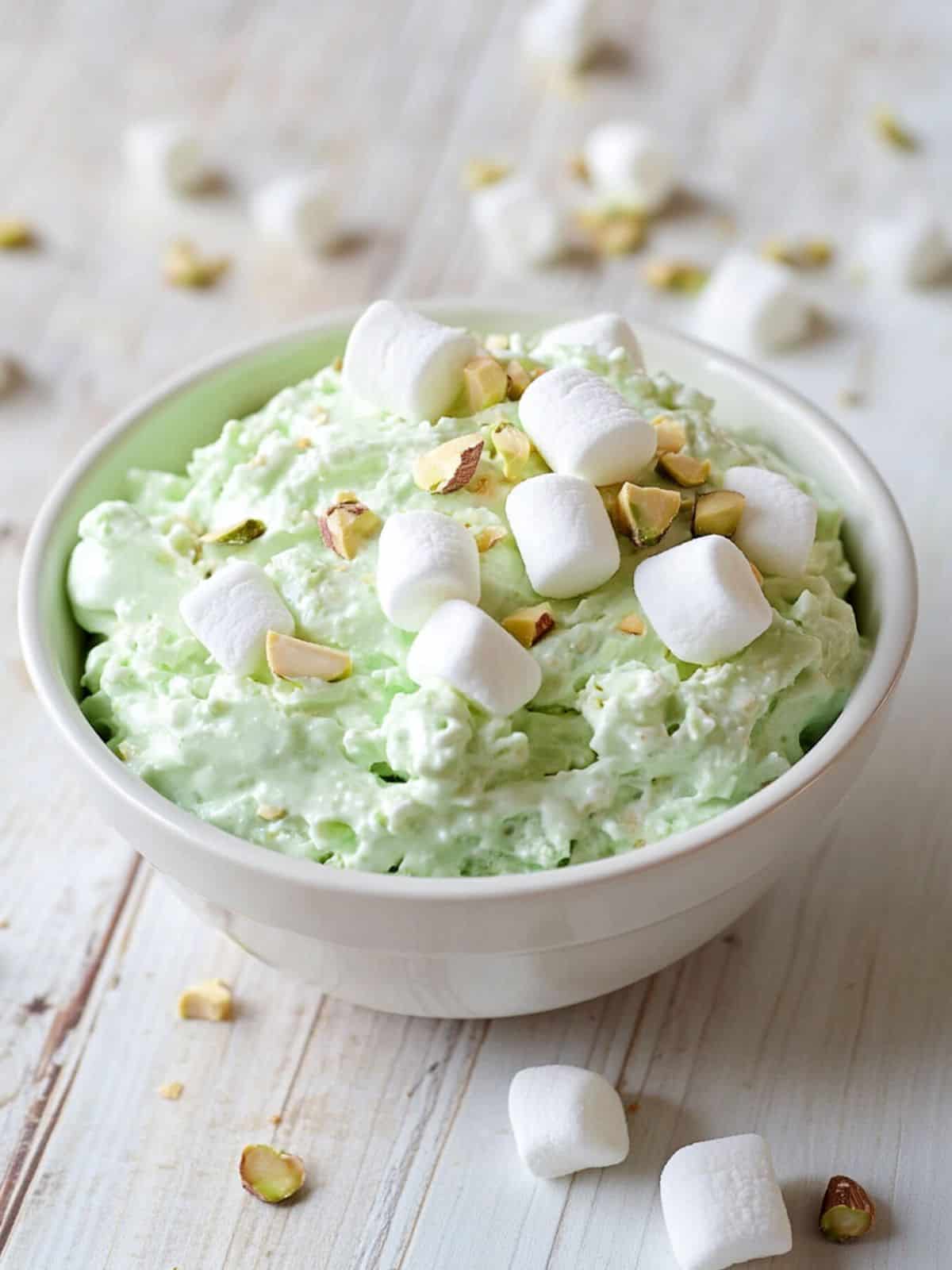 An image of watergate salad in a white bowl on a white painted table. On the table are marshmallows and pistachio nibs.