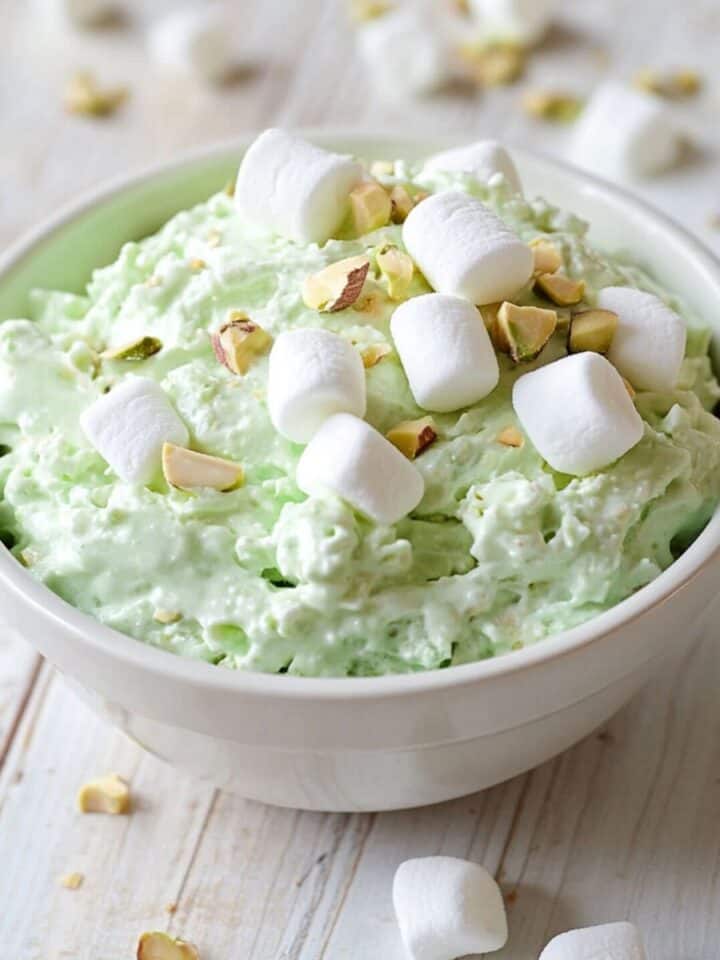 An image of watergate salad in a white bowl on a white painted table. On the table are marshmallows and pistachio nibs.