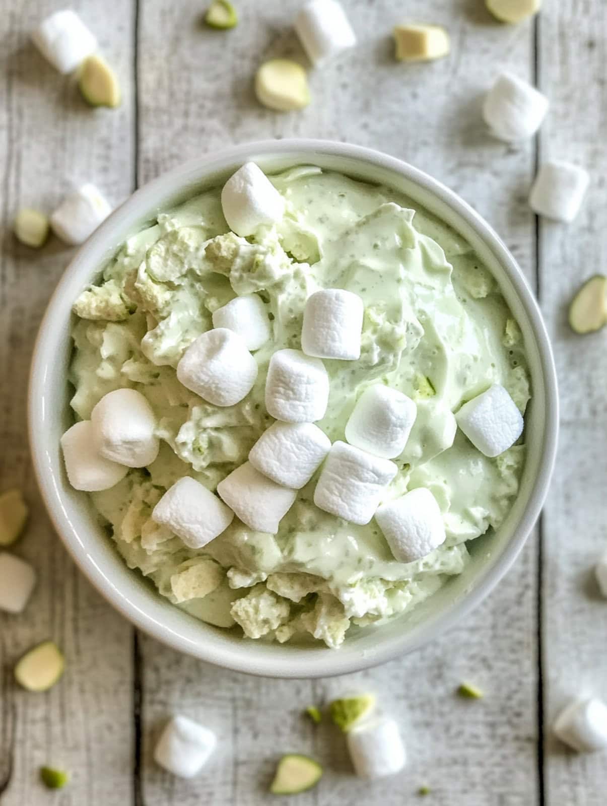 An image of watergate salad in a white bowl on a white painted table. On the table are marshmallows and pistachio nibs.